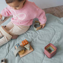 A toddler engages in sensory play on a mat with the Montessori First Birthday Gift Set – 3 Toys, featuring a wooden shape-sorting toy, a set of nesting boxes, and a curved stacking toy.