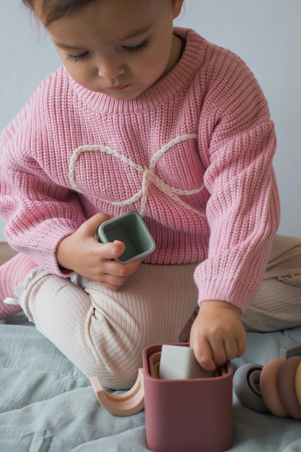 A young child in a pink sweater engages in sensory play with a Stack Happy Trio – Montessori Stacking Set of nested, pastel-colored silicone stackers.
