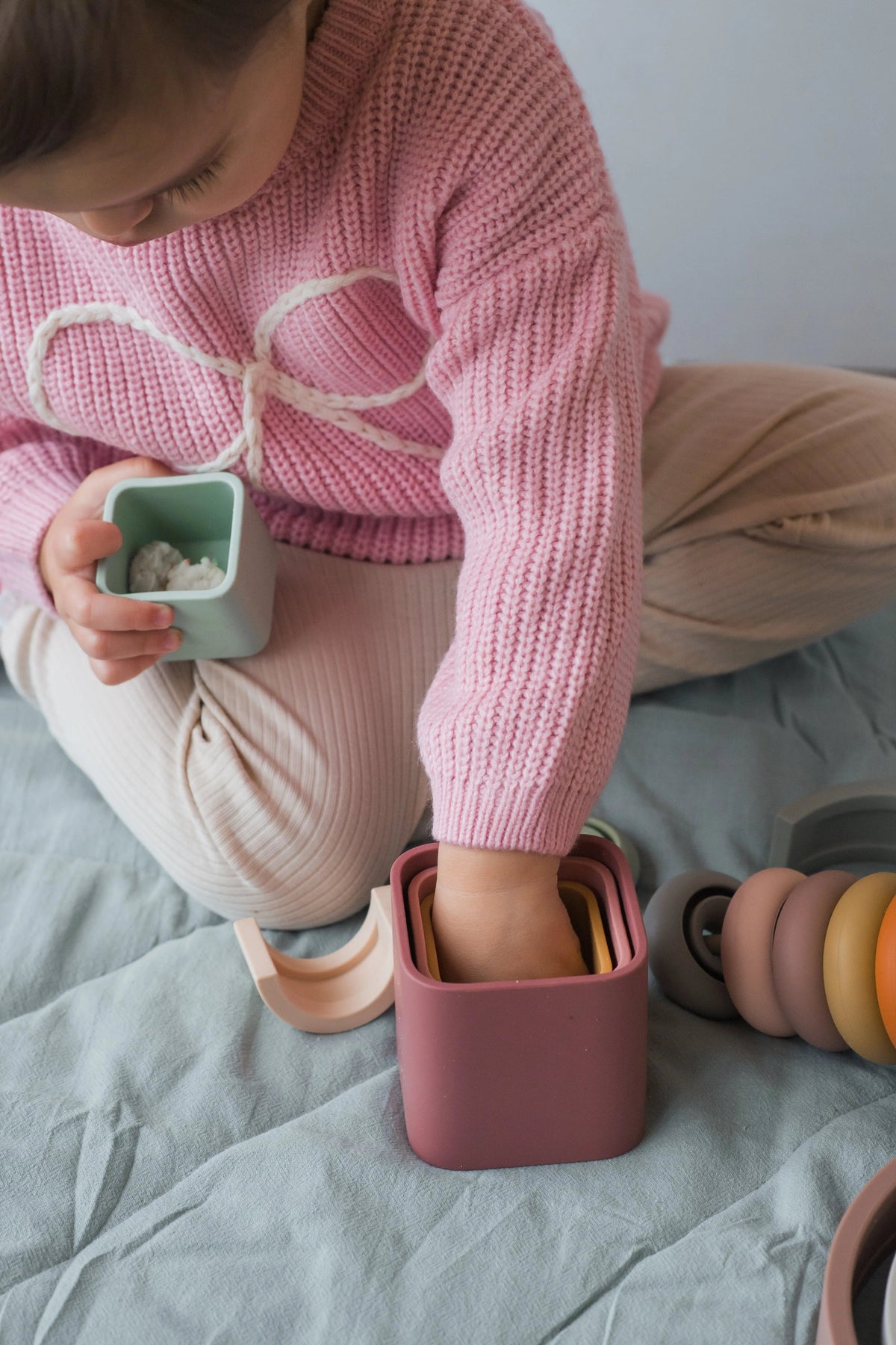 A child sits on a bed engaging in sensory play with a Stack Happy Trio – Montessori Stacking Set of nested silicone blocks and stacking toys.
