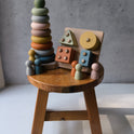 A Montessori Sensory Starter Kit – 3 Toy Set featuring a wooden stacking ring tower, a shape-sorting puzzle board, and several small wooden peg dolls arranged on a small wooden stool to help develop fine motor skills.