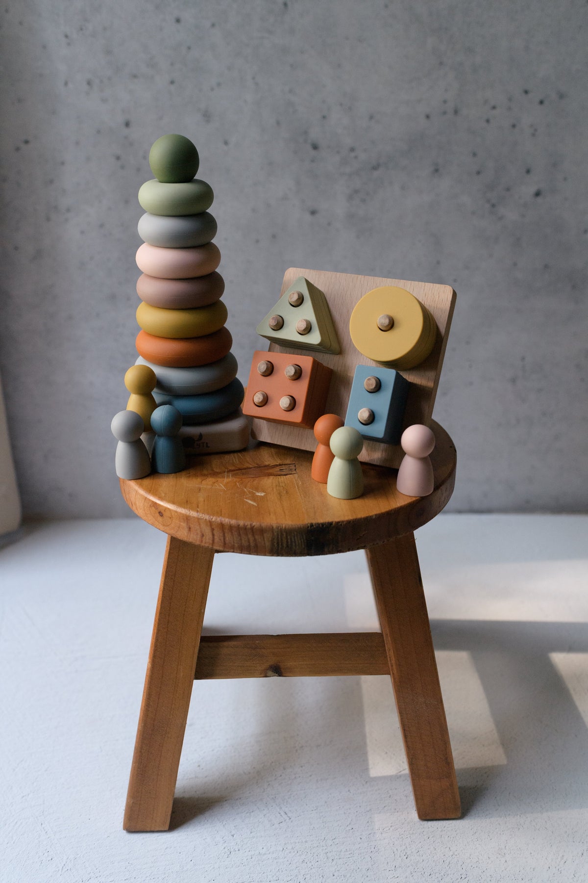 A Montessori Sensory Starter Kit – 3 Toy Set featuring a wooden stacking ring tower, a shape-sorting puzzle board, and several small wooden peg dolls arranged on a small wooden stool to help develop fine motor skills.