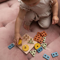 A toddler practices fine motor skills by placing a yellow circular piece onto a Montessori Wood & Silicone Shape Puzzle, surrounded by various geometric wooden and silicone shapes.