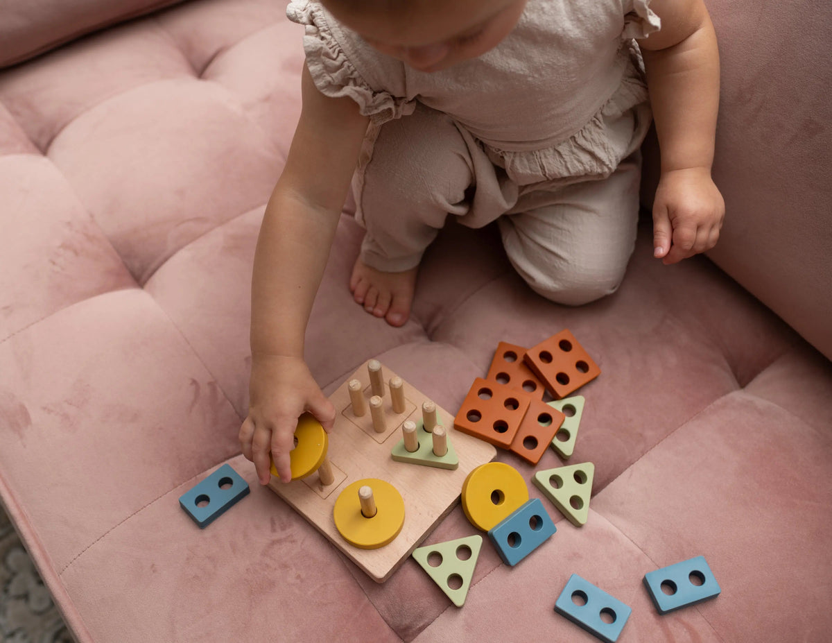 A toddler practices fine motor skills by placing a yellow circular piece onto a Montessori Wood & Silicone Shape Puzzle, surrounded by various geometric wooden and silicone shapes.