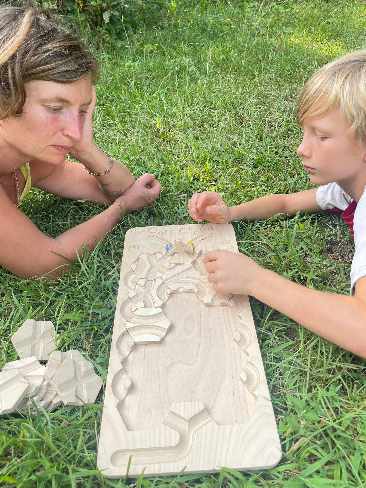 An adult and a child sitting in the grass, assembling The Oort Cloud – Wooden Marble Run Puzzle on a board.
