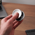 A hand spins the ONO Slider Aluminum, a round silver calming fidget toy with magnetized disks, on a wooden surface near a laptop and smartphone.