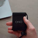 A hand holds a black pouch labeled ONO, then pulls out a compact ONO Slider Aluminum foldable magnifying glass and opens it to reveal its circular lens, with a wooden surface and part of a laptop in the background.