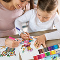 A woman and a child color owl-themed drawings at a table with the Vibrant Dual Tip Brush Pen Marker Set. The child uses pink while the woman chooses purple, and an extensive range of markers from 12 to 160 colors enhances their vibrant creative session.