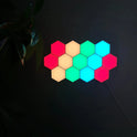 Twelve Hexagonal LED Modular Night Light Panels for Sensory Rooms, glowing in red, yellow, and blue-green, are mounted on a dark wall with a cable at the bottom right and leaves partly visible on the left.