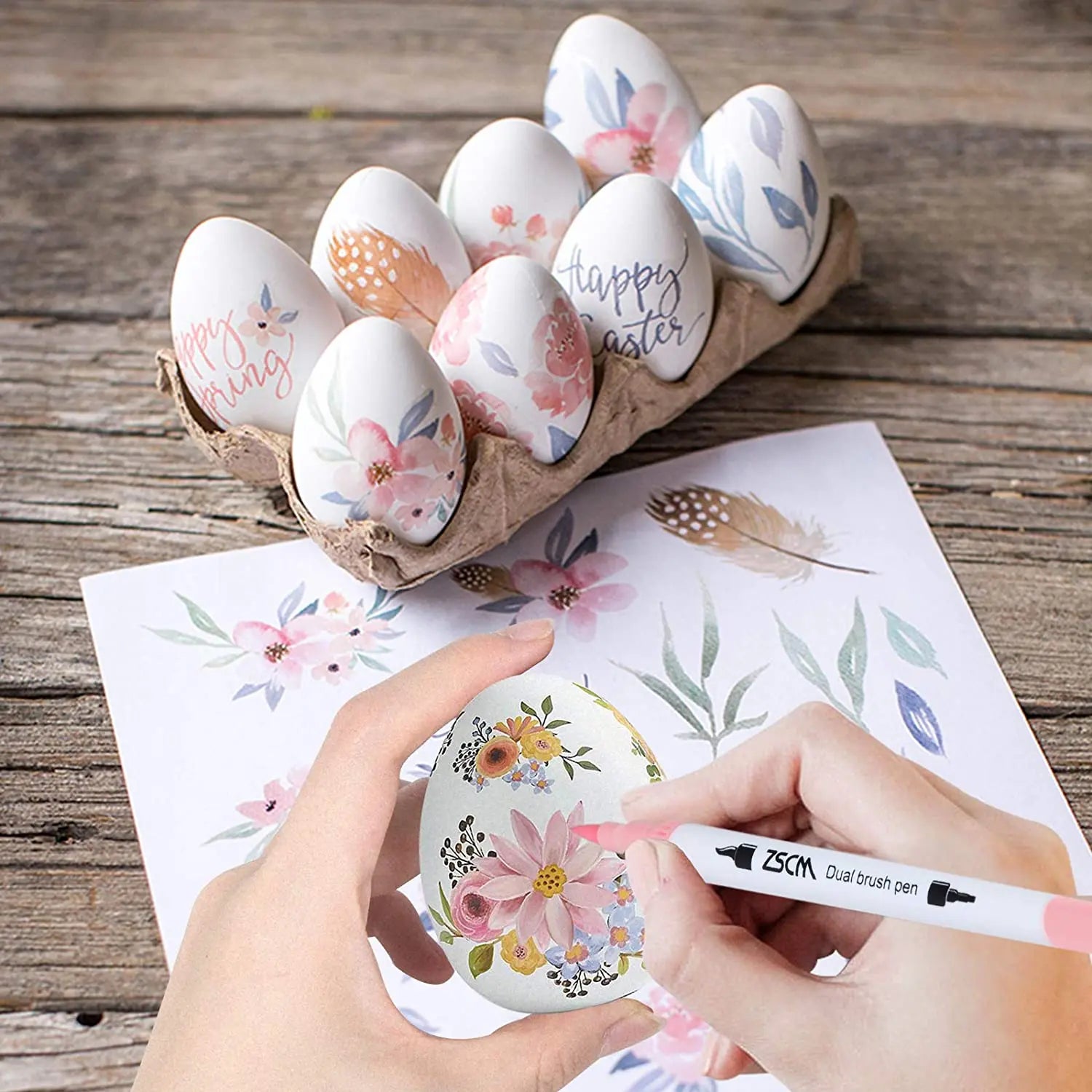 Using the Vibrant Dual Tip Brush Pen Marker Set (12 to 160 colors), a person decorates a white Easter egg with floral designs on floral-patterned paper. Nearby, a carton holds similarly adorned eggs with Happy Easter messages, showcasing the markers versatility and extensive color range.