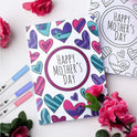 A Mothers Day card with vibrant hearts is surrounded by pink flowers and the Vibrant Dual Tip Brush Pen Marker Set, which offers 12 to 160 colors for creative drawing, coloring, and sketching.