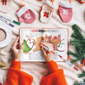 A person in an orange sweater decorates a journal page with a Christmas gingerbread recipe using the Vibrant Dual Tip Brush Pen Marker Set. They are surrounded by festive items like small stockings, a pine branch, and a cup of coffee on a cozy blanket.