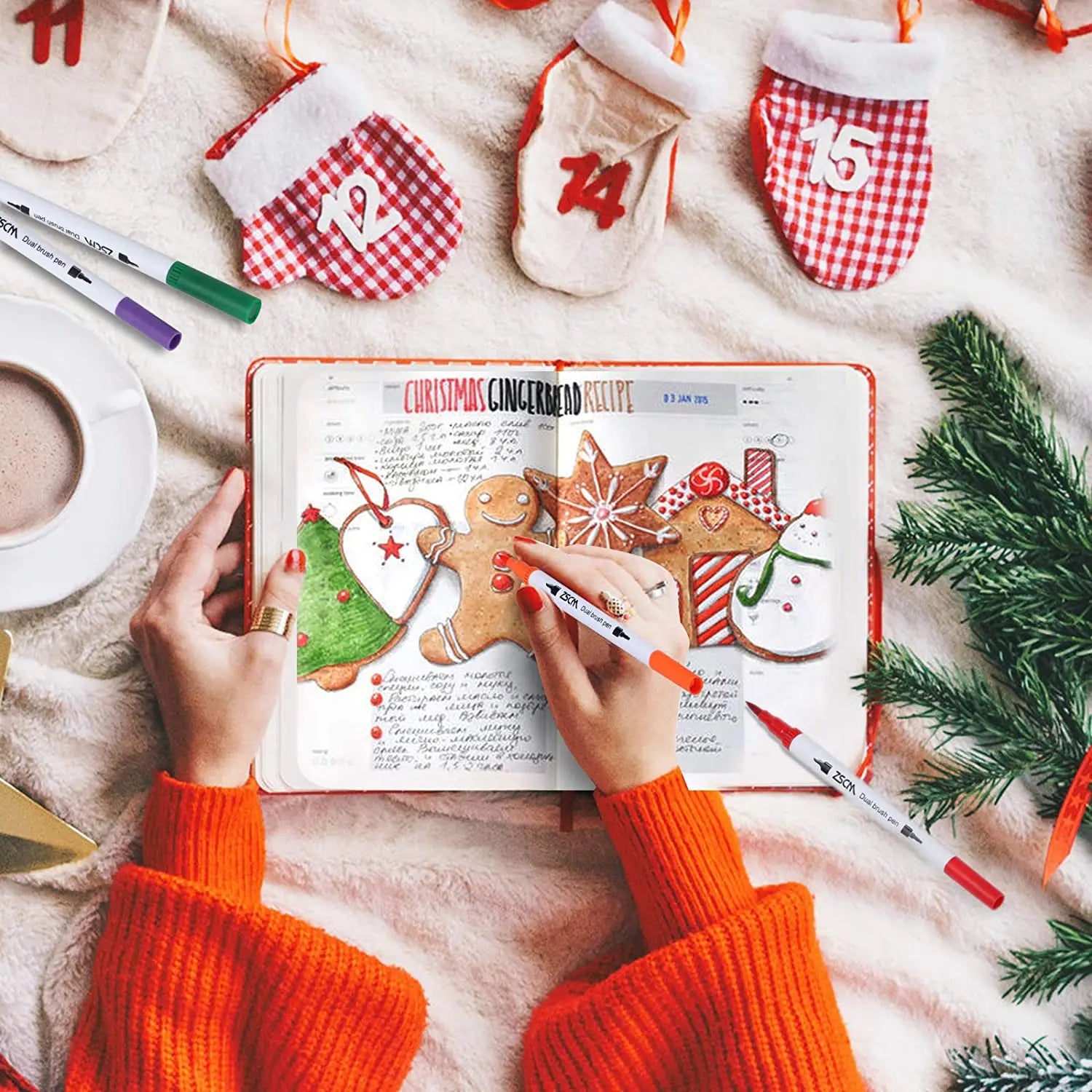 A person in an orange sweater decorates a journal page with a Christmas gingerbread recipe using the Vibrant Dual Tip Brush Pen Marker Set. They are surrounded by festive items like small stockings, a pine branch, and a cup of coffee on a cozy blanket.