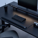 A minimalist black desk setup features the Desk Mat Bundle, including a sleek vegan leather mat, keyboard, large monitor, black cup with straw, camera, and ergonomic wrist cushion, all neatly arranged beside an office chair.
