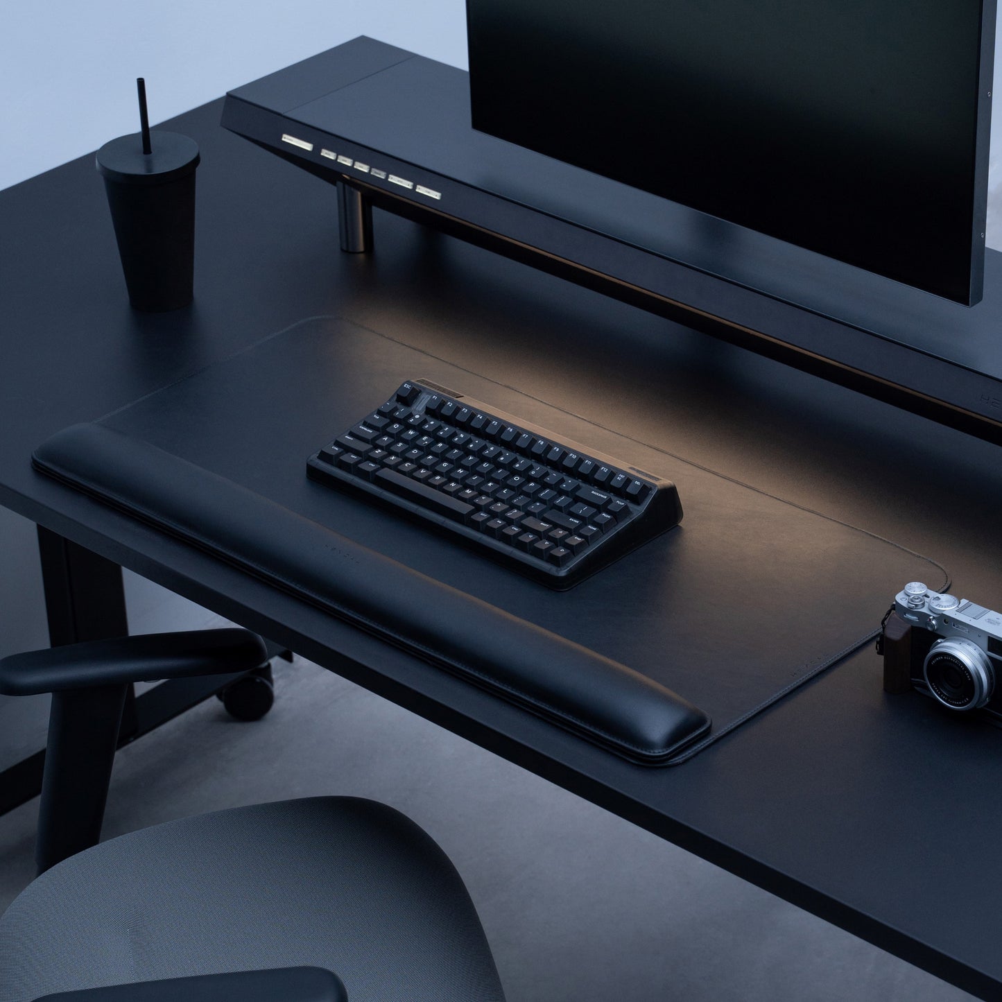A minimalist black desk setup features the Desk Mat Bundle, including a sleek vegan leather mat, keyboard, large monitor, black cup with straw, camera, and ergonomic wrist cushion, all neatly arranged beside an office chair.
