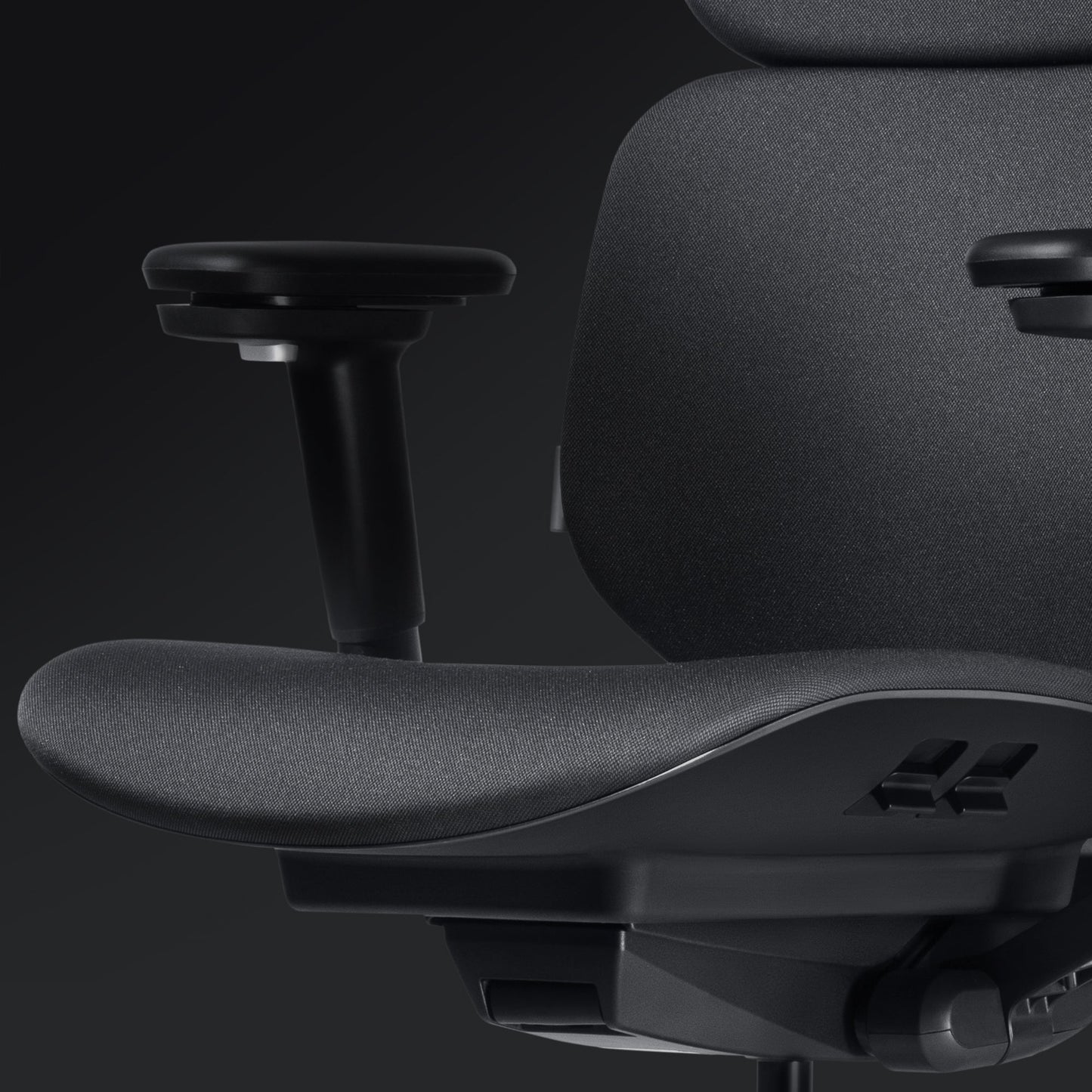 Close-up of the Hexcal Inspire Chair, featuring dark grey fabric upholstery, adjustable armrest, seat, and part of the backrest, set against a dark background.