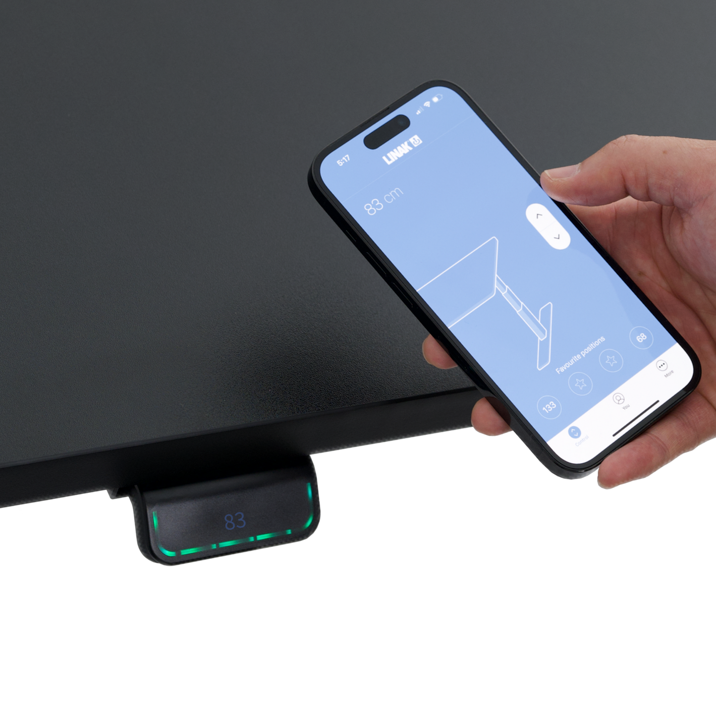 A hand holds a smartphone with an app for adjusting the Hexcal Elevate Standing Desk, near the desk’s digital control panel displaying a height of 83 cm.
