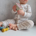 A baby sits on the floor holding a small toy box with six smooth, colorful Silicone Mini Peg Dolls (Set of 6) scattered nearby for pretend play.