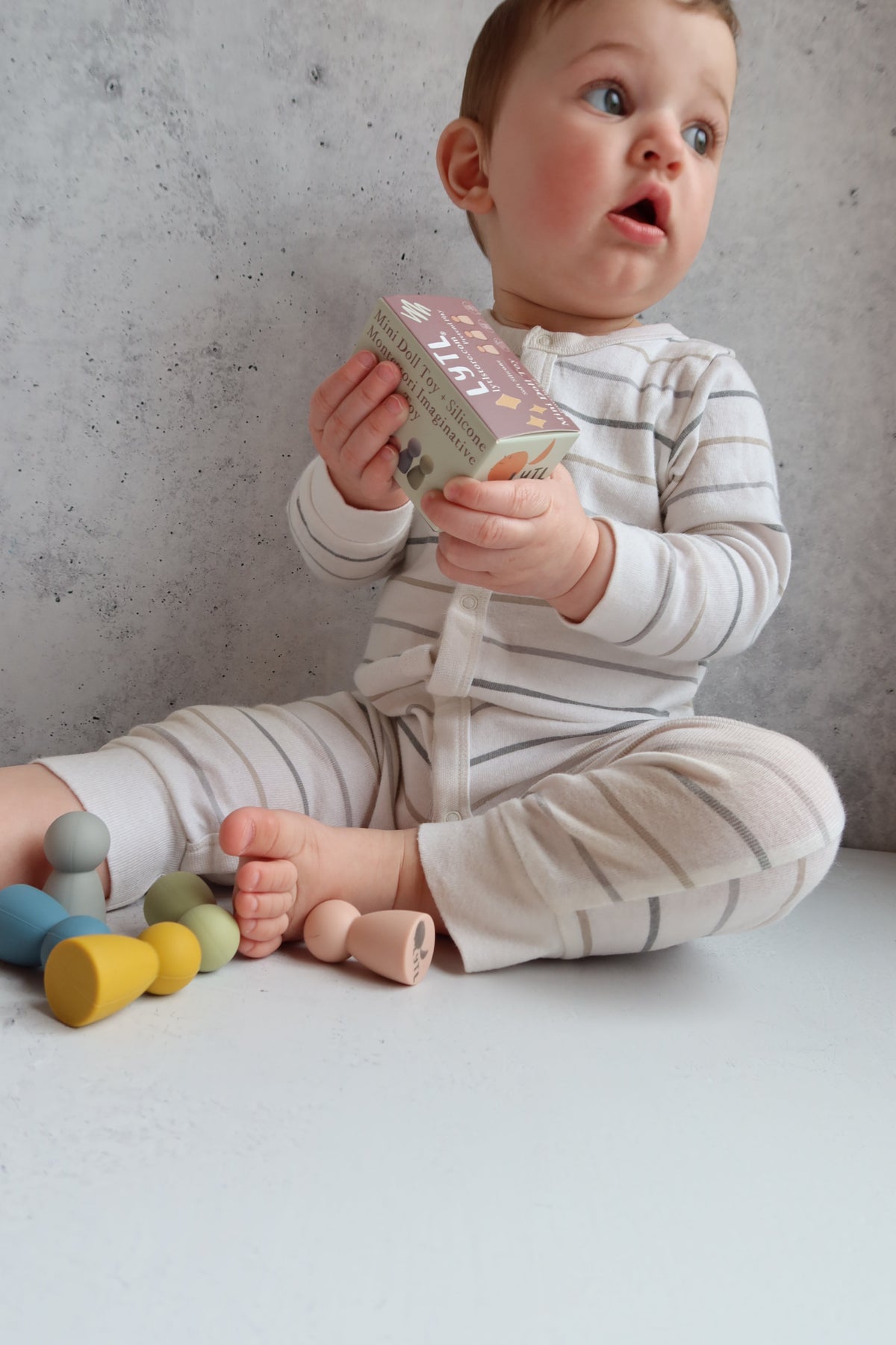 A baby sits on the floor holding a small toy box with six smooth, colorful Silicone Mini Peg Dolls (Set of 6) scattered nearby for pretend play.