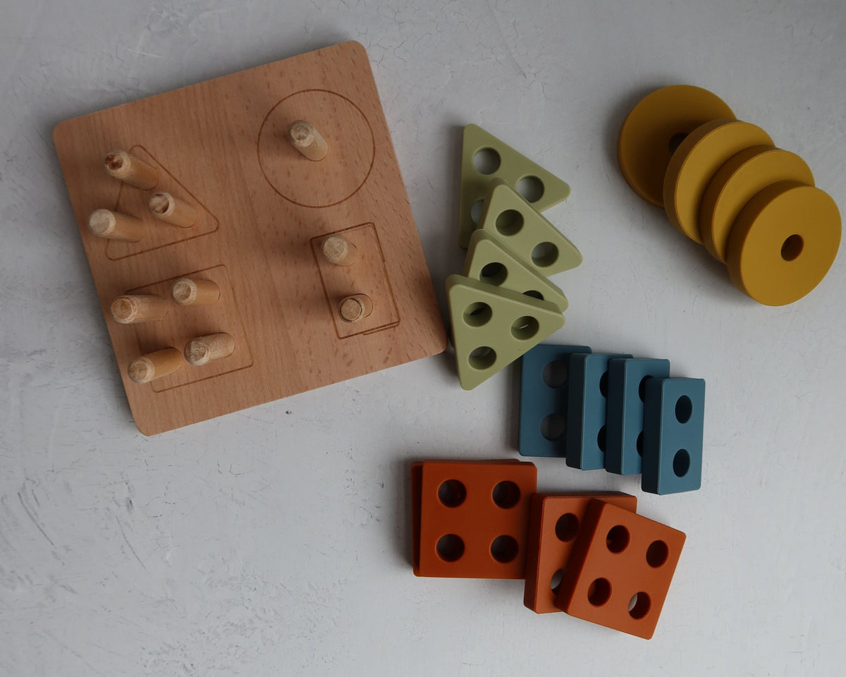 The Montessori Wood & Silicone Shape Puzzle is a wooden shape sorter with pegs next to groups of yellow circles, olive triangles, blue rectangles, and rust-colored squares, designed to help children develop fine motor skills.