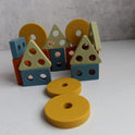 Blue, orange, yellow, and green geometric blocks from the Montessori Wood & Silicone Shape Puzzle arranged to resemble house shapes, with two yellow circular discs from this silicone baby toy resting in the foreground.