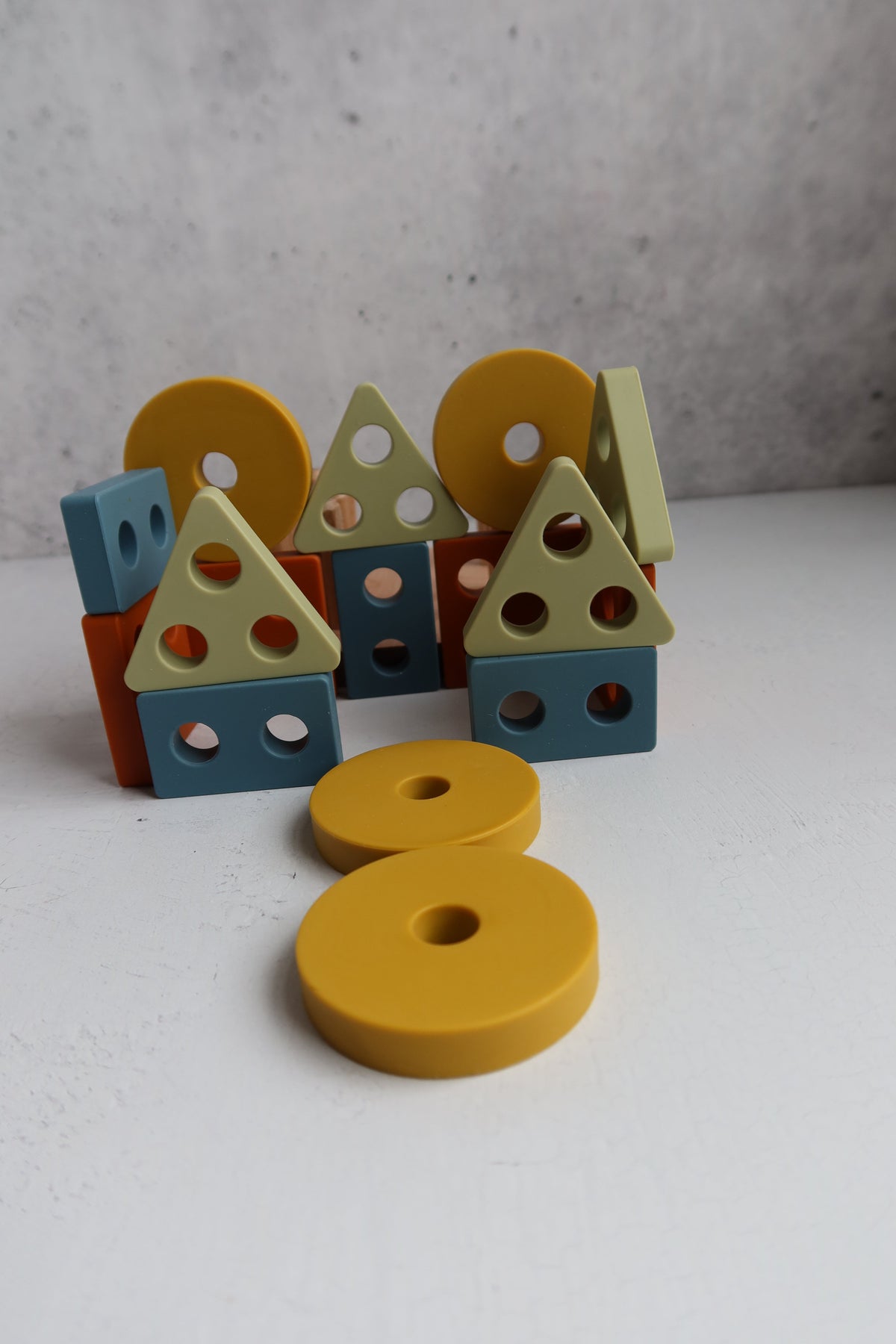 Blue, orange, yellow, and green geometric blocks from the Montessori Wood & Silicone Shape Puzzle arranged to resemble house shapes, with two yellow circular discs from this silicone baby toy resting in the foreground.