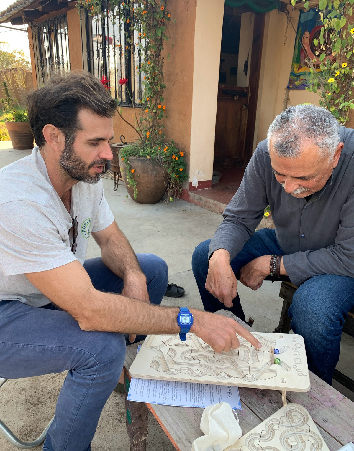 Two men sit at an outdoor table playing with The Oort Cloud – Wooden Marble Run Puzzle.