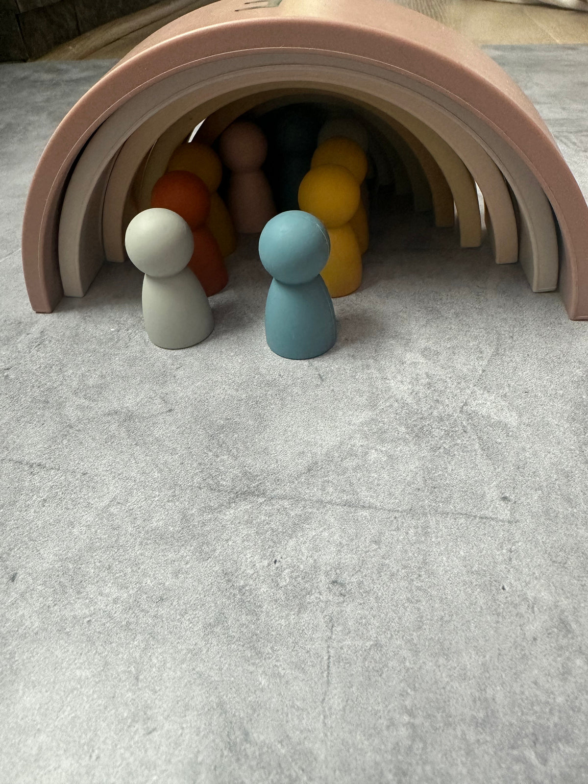 Colorful, minimalist Silicone Mini Peg Dolls (Set of 6) arranged in front of and underneath a nested, arched stacker toy, perfect for pretend play.
