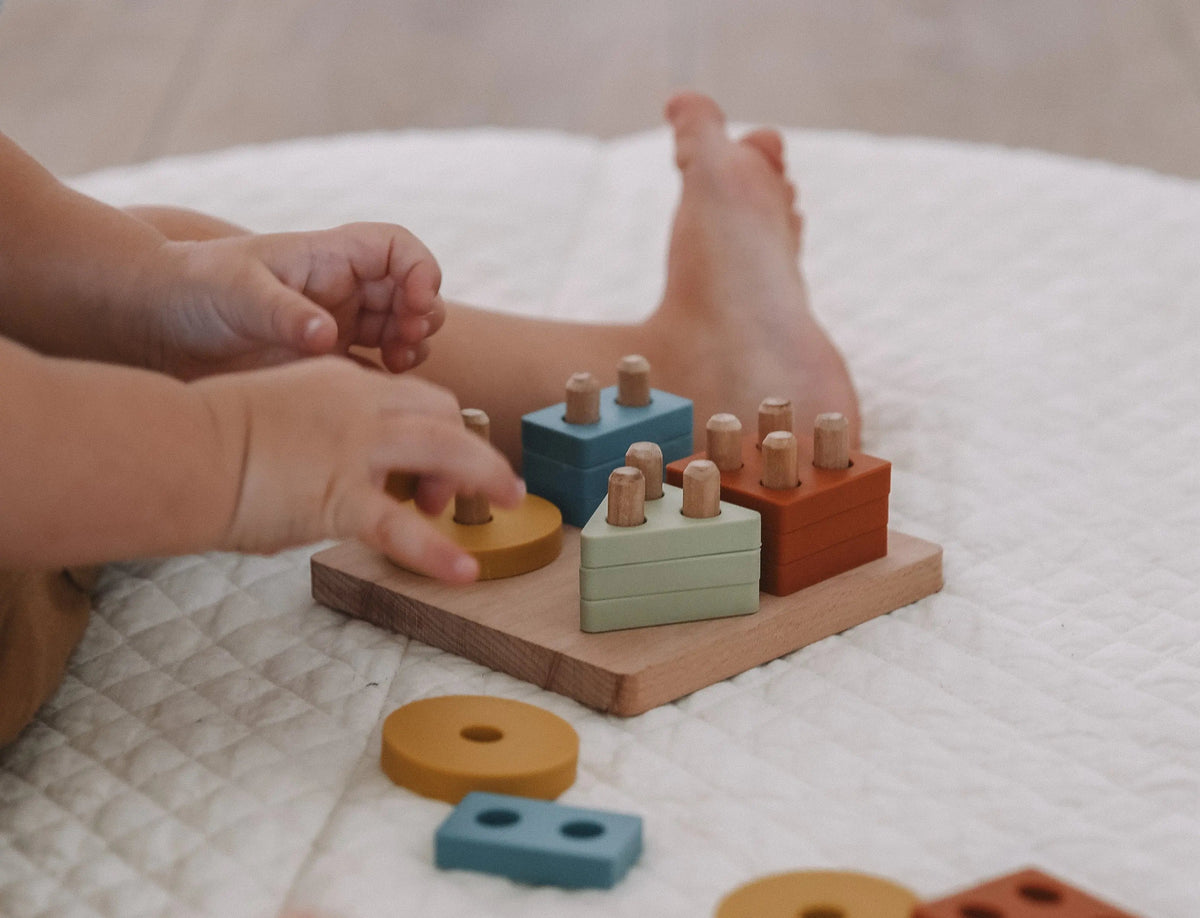 A child’s hands play with a Montessori Wood & Silicone Shape Puzzle, using blue, yellow, green, and orange wooden geometric shapes to build fine motor skills.