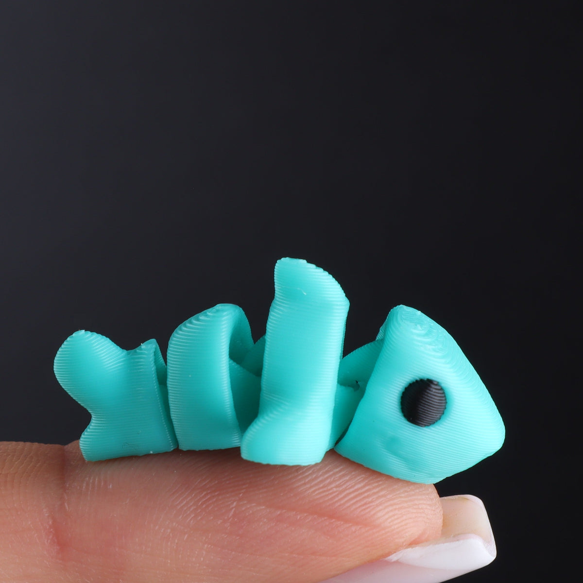 A small, turquoise articulated fish from the Micro Animal Fidgets – Surprise Pack, perfect as micro 3D-printed desk decor, balanced on a person’s fingertip.