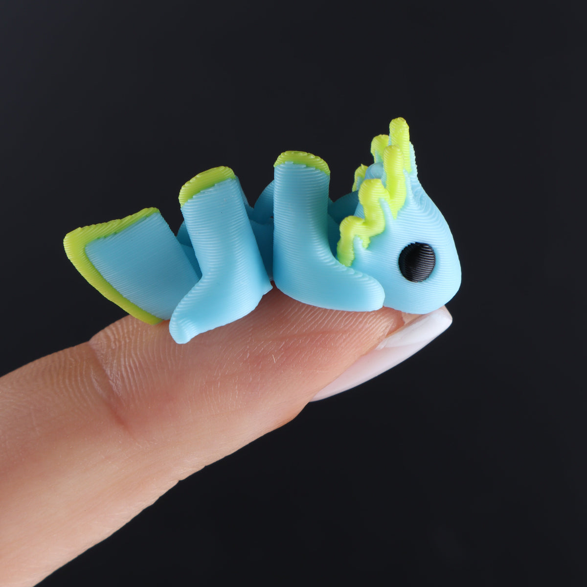 A tiny, blue, articulated 3D-printed axolotl with yellow accents from a Micro Animal Fidgets – Surprise Pack balanced on a person’s fingertip as mini desk decor.