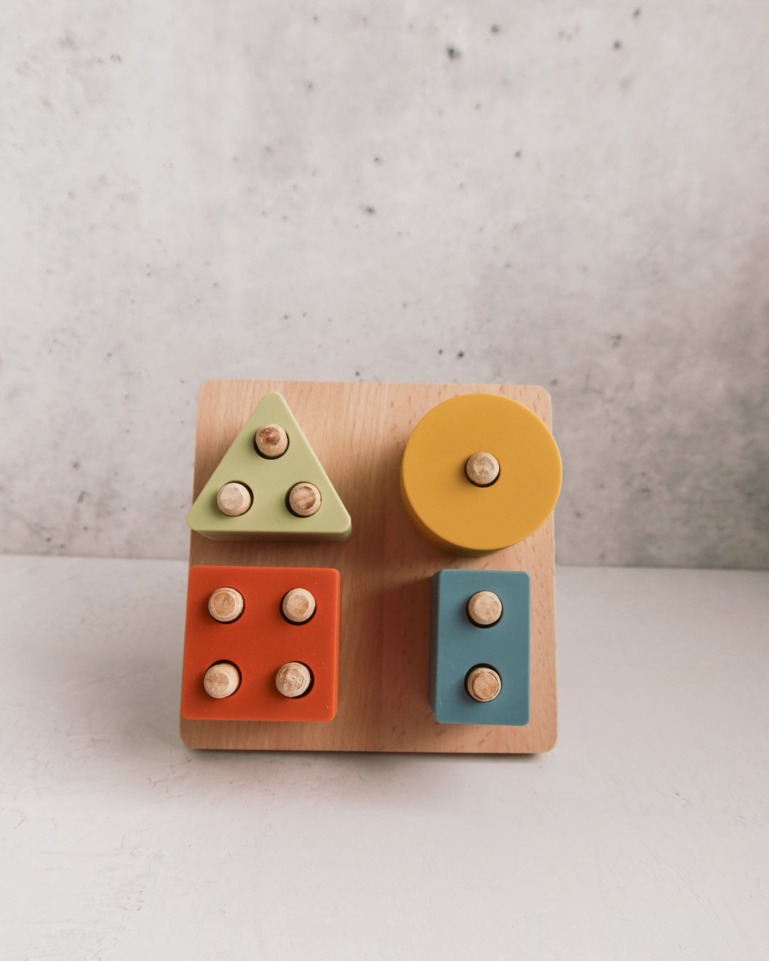 A Montessori Wood & Silicone Shape Puzzle featuring a square wooden board with green, yellow, orange, and blue geometric pieces, each with a small wooden peg for fine motor skills.