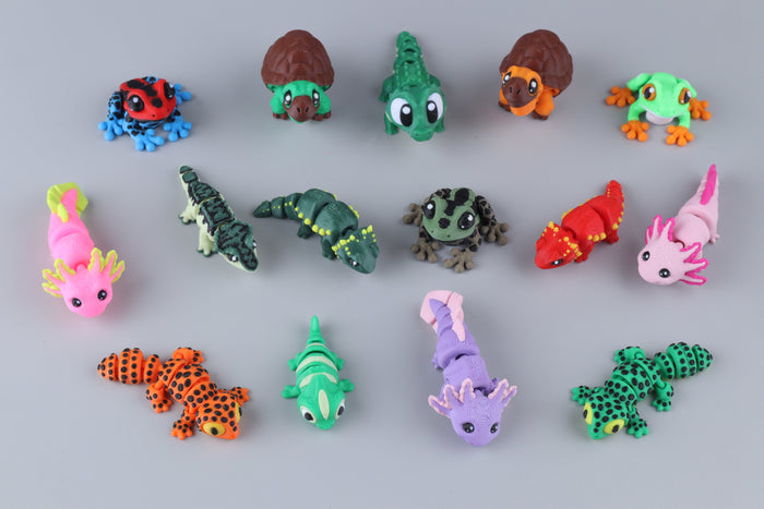 Fifteen colorful 3D-printed Mini Reptile & Amphibian Fidgets – 15 Pack, including frogs, turtles, crocodiles, and axolotls, arranged on a gray surface.