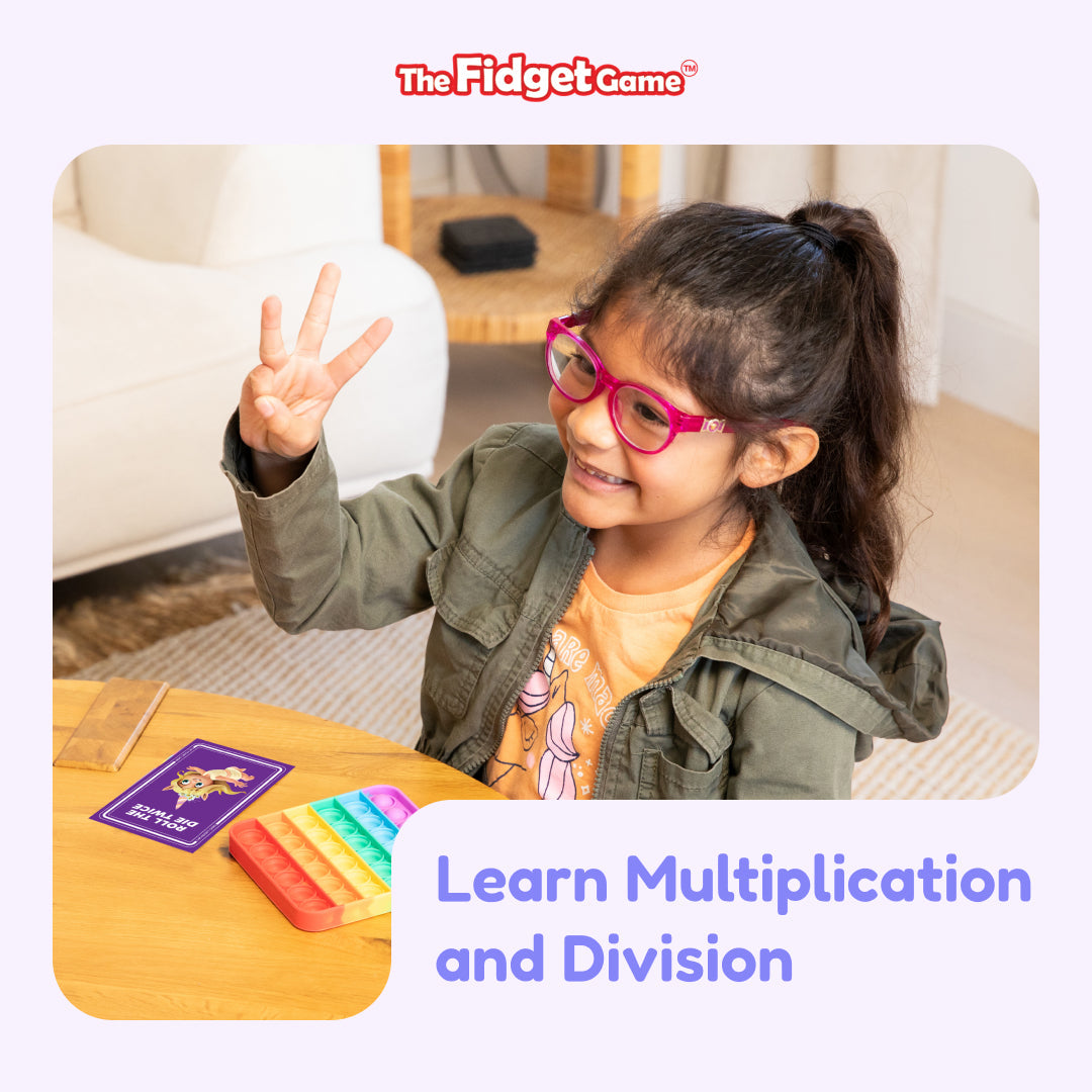 A young girl holds up three fingers while sitting at a table with a Math Card Pack Bundle (Multiplication + Division) and a rainbow-colored fidget toy.