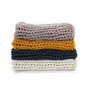 A stack of four Knit Weighted Blankets—super soft in light gray on top, then mustard yellow, dark navy blue, and cream below—all neatly folded against a white background.
