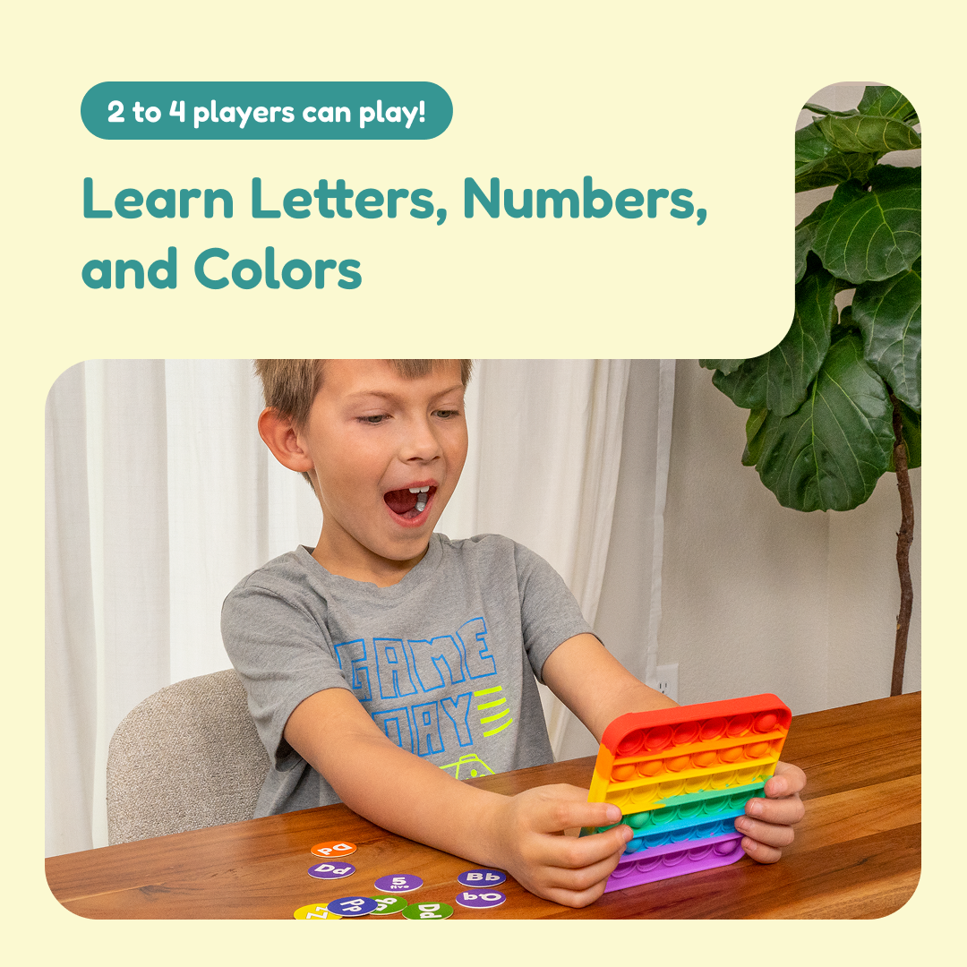A child excitedly holds a rainbow-colored fidget toy at a table scattered with alphabet and number discs from the My First Learning Game Bundle, which teaches letters, numbers, and colors to 2 to 4 players.