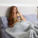 A woman with long brown hair smiles while sitting up in bed, holding a mug. She is wrapped in a chunky light gray knit blanket, and the bed is dressed in Forever Chill Bamboo Sheets with a light-colored headboard.