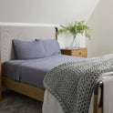 A neatly made bed with Forever Chill Bamboo Sheets in Arctic Grey, a chunky knit blanket, and a wooden nightstand holding a clear vase with green foliage, set in a cozy, softly lit bedroom.