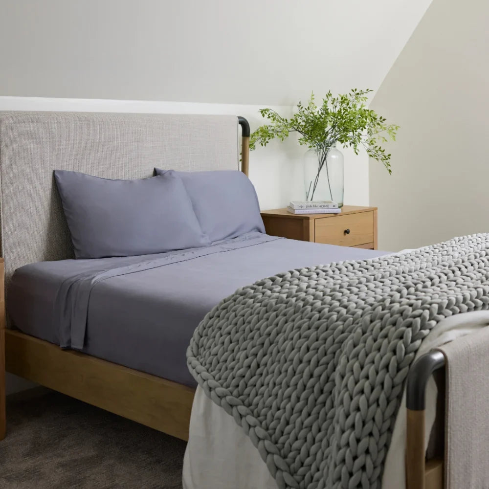 A neatly made bed with Forever Chill Bamboo Sheets in Arctic Grey, a chunky knit blanket, and a wooden nightstand holding a clear vase with green foliage, set in a cozy, softly lit bedroom.