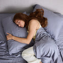 A woman with long red hair sleeps on her side in bed, hugging a pillow and covered with Forever Chill Bamboo Sheets in gray, while wearing a light-colored pajama set.