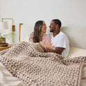 A smiling couple sits up in bed, wrapped in the Ice Knit Weighted Blanket, enjoying a cozy moment together in a softly lit bedroom.