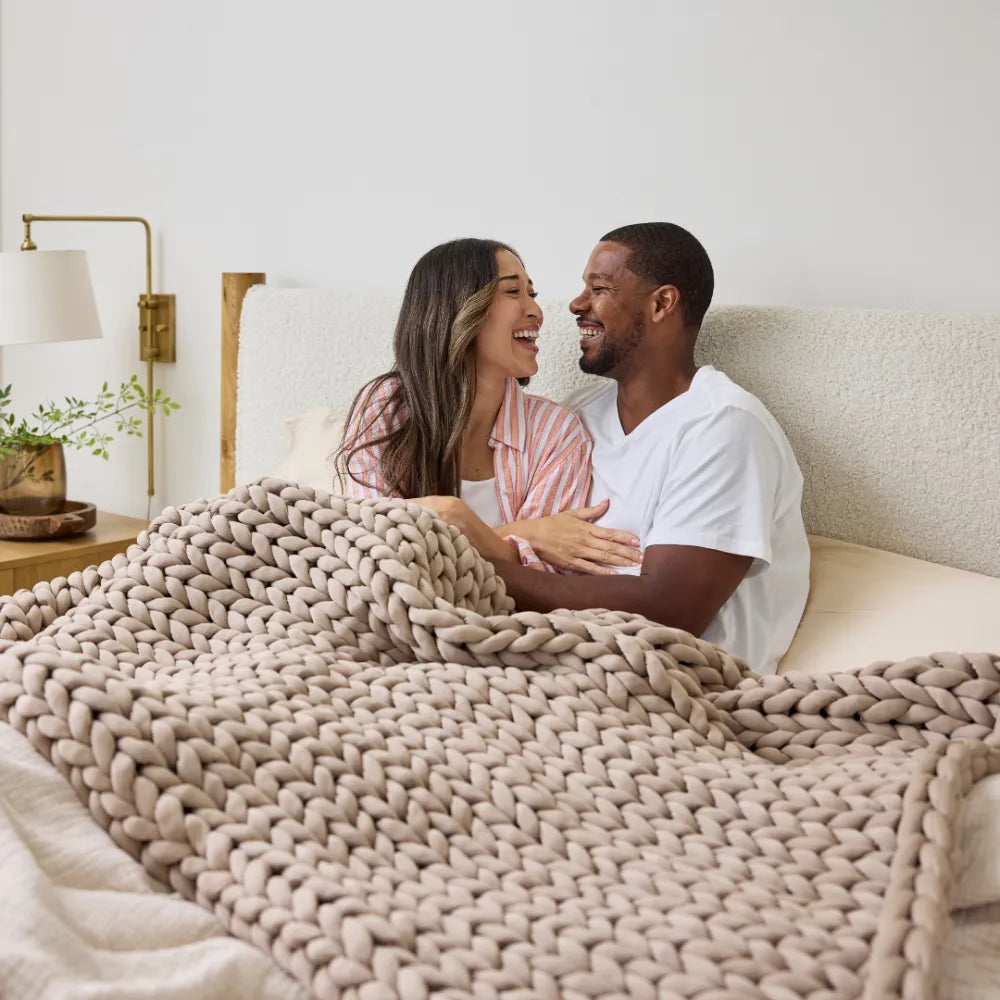 A smiling couple sits up in bed, wrapped in the Ice Knit Weighted Blanket, enjoying a cozy moment together in a softly lit bedroom.