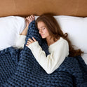 A woman with long brown hair, wearing a white long-sleeve shirt, lies peacefully on a bed with her eyes closed, hugging the Ice Knit Weighted Blanket—a breathable, chunky knit navy blue weighted blanket.