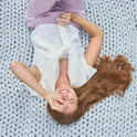 A smiling woman with long brown hair lies on the Ice Knit Weighted Blanket, wearing light purple pants and a white top with a sweater over her shoulders, making a peace sign over her eye.