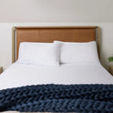 A neatly made bed with a tan leather headboard, two white pillows, and crisp Snow White Forever Chill Bamboo Sheets, topped with a chunky dark blue knit blanket at the foot. A small green plant sits on the bedside table.