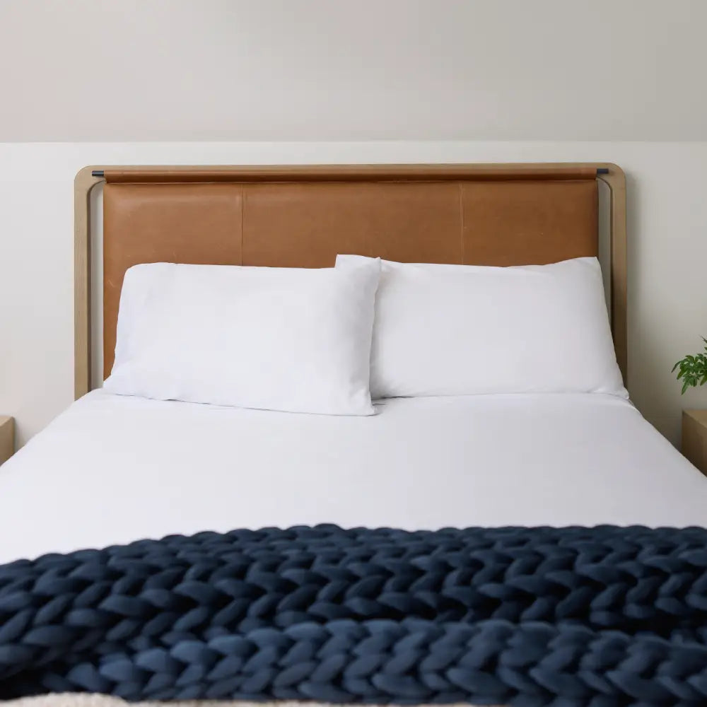 A neatly made bed with a tan leather headboard, two white pillows, and crisp Snow White Forever Chill Bamboo Sheets, topped with a chunky dark blue knit blanket at the foot. A small green plant sits on the bedside table.