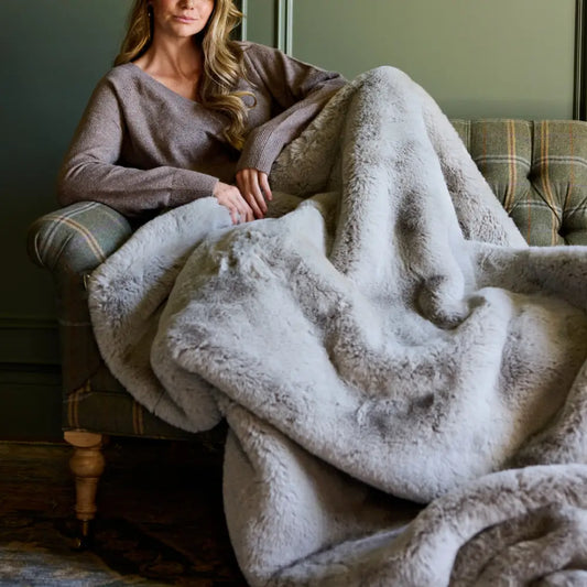 A woman with long, wavy hair sits on a plaid armchair with the Snuggle Luxe Blanket over her legs. Dressed in a brown sweater, she relaxes in a cozy room with muted green walls that enhance the inviting atmosphere.