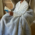 A person in a white shirt sits in a chair, wrapped in the plush Snuggle Luxe Blanket, reading a blue-covered book in a cozy, well-lit room.