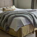 A softly lit bedroom features a cozy bed with a white Snuggle Luxe Blanket, a fluffy gray throw, a decorative pillow, and a beige bed skirt.
