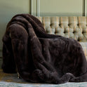 A large mocha Snuggle Luxe Blanket draped over a green and yellow plaid upholstered sofa, set against a muted green paneled wall.