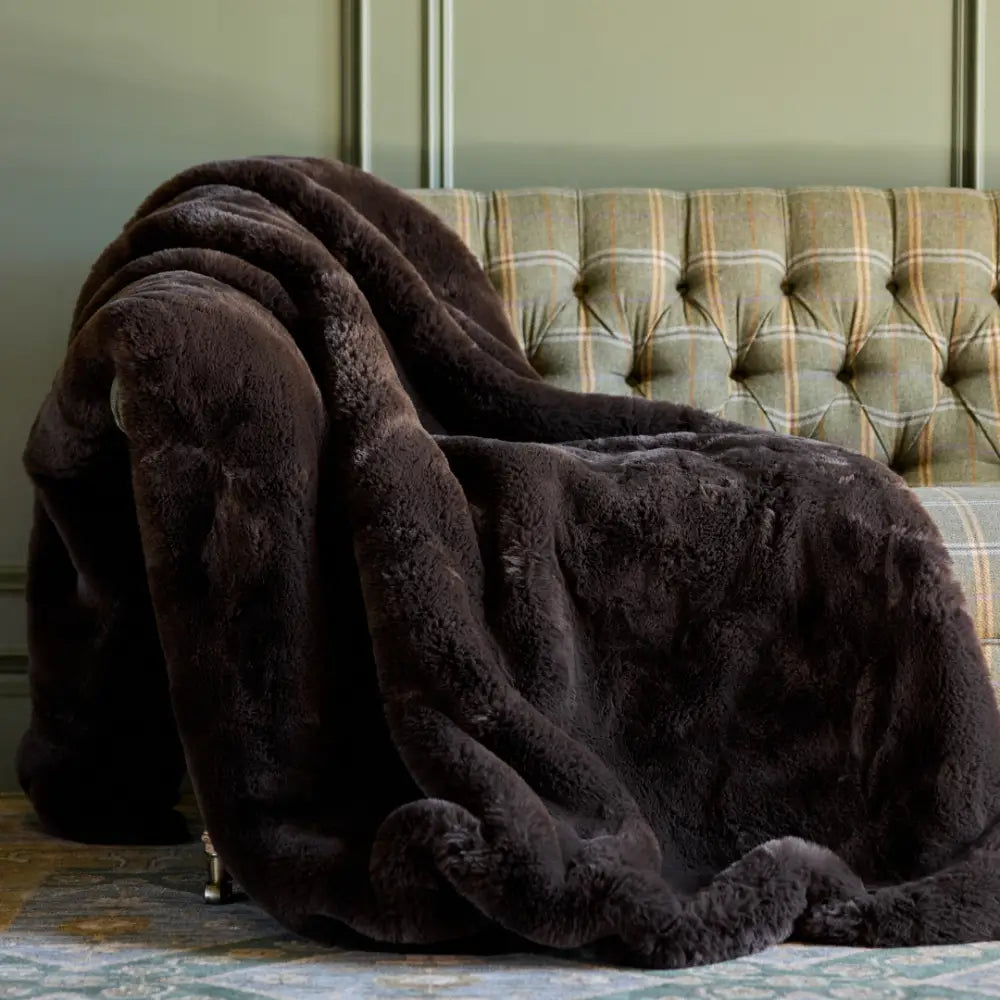 A large mocha Snuggle Luxe Blanket draped over a green and yellow plaid upholstered sofa, set against a muted green paneled wall.